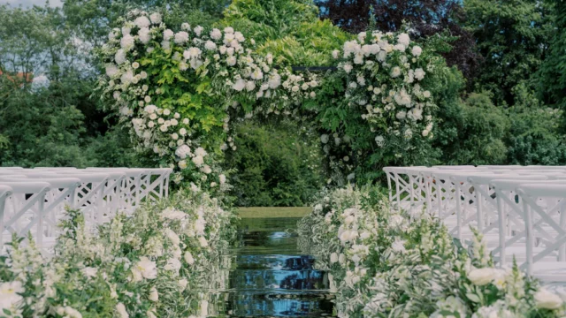 Florist: Larry Walshe. Wedding wise, glass, water, green and white foliage wedding arch, white chairs.